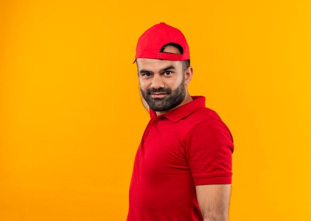 Livreur barbu en uniforme rouge et chapeau souriant confiant debout sur mur orange