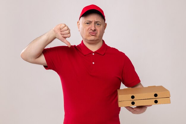 Livreur insatisfait en uniforme rouge et casquette regardant la caméra montrant les pouces vers le bas tenant des boîtes à pizza debout sur fond blanc