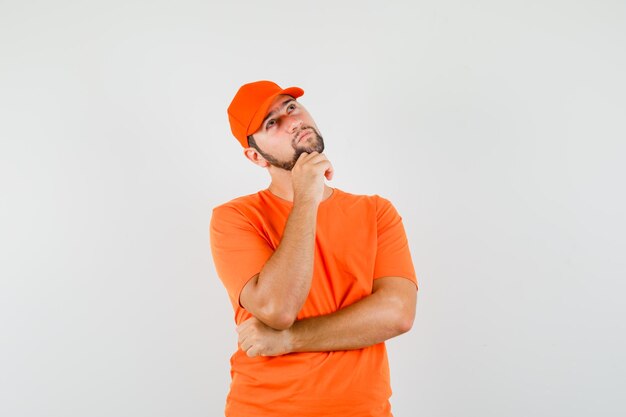 Livreur en levant la main sur le menton en t-shirt orange, casquette et pensif. vue de face.