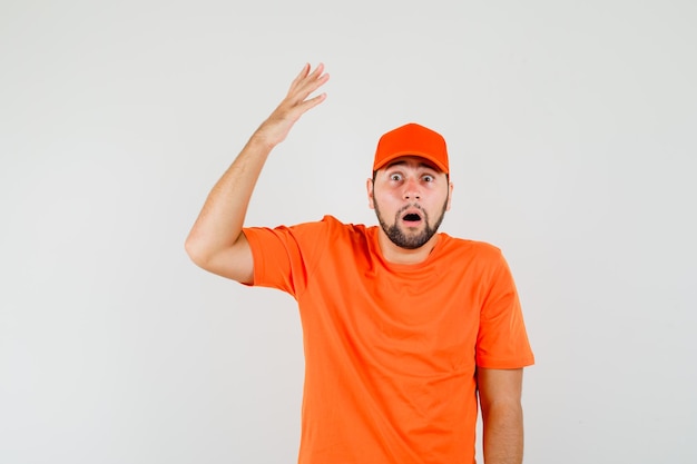 Photo gratuite livreur levant la main en t-shirt orange, casquette et l'air choqué. vue de face.