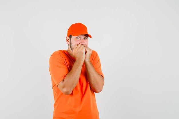 Livreur mordant émotionnellement les poings en t-shirt orange, casquette et ayant l'air effrayé, vue de face.