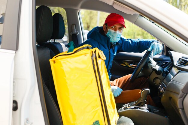 Livreur de nourriture dans un masque de protection et des gants avec un sac à dos thermo près d'une voiture pendant la période de quarantaine