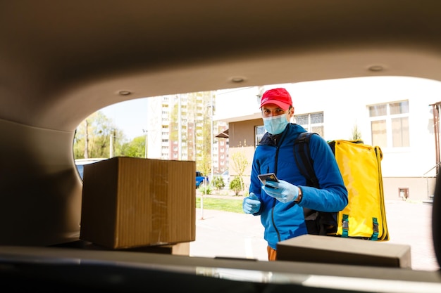 Livreur de nourriture dans un masque de protection et des gants avec un sac à dos thermo près d'une voiture pendant la période de quarantaine