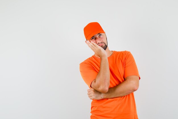 Livreur penché la joue sur la paume surélevée en t-shirt orange, casquette et l'air pensif. vue de face.