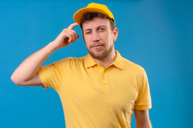 Livreur en polo jaune et casquette ayant des doutes tout en se grattant la tête un clin de œil debout sur bleu isolé