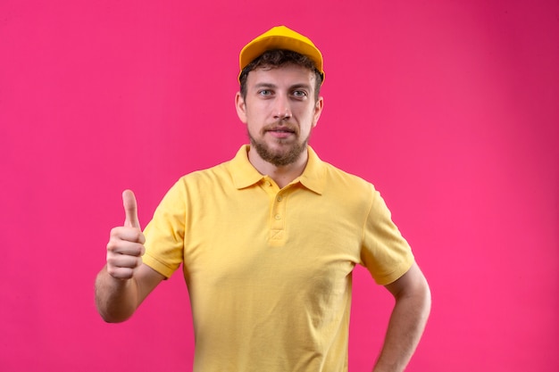 Photo gratuite livreur en polo jaune et casquette à la confiance montrant les pouces vers le haut debout sur rose isolé