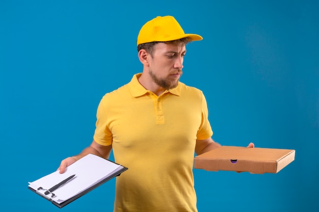 Livreur en polo jaune et casquette debout avec boîte à pizza et presse-papiers à la main en fronçant les sourcils sur bleu