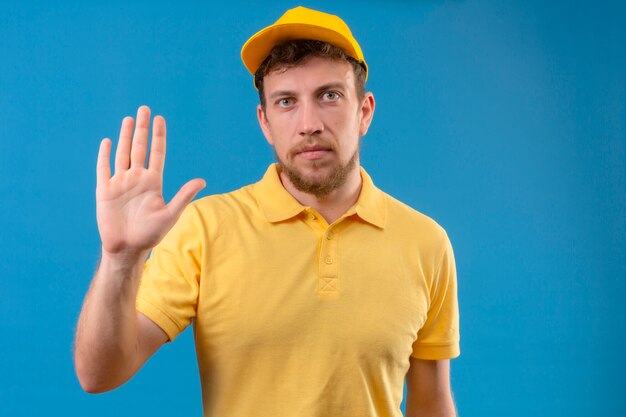 Livreur en polo jaune et casquette debout avec la main ouverte faisant panneau d'arrêt avec un geste de défense d'expression sérieuse et confiante sur bleu isolé