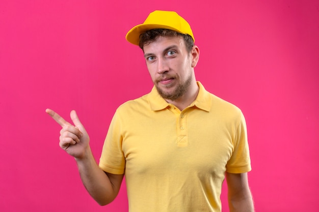 Livreur en polo jaune et casquette pointant avec l'index sur le côté avec sourire confiant sur rose isolé