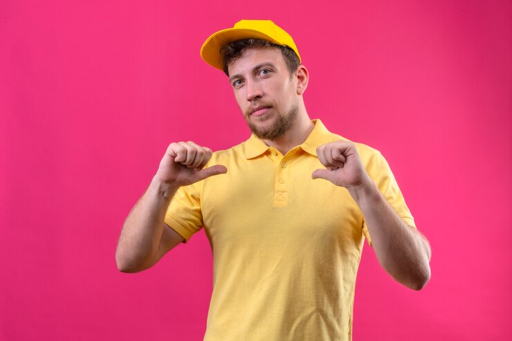 Photo gratuit livreur en polo jaune et casquette pointant vers lui-même avec les deux pouces à la confiance de soi-même debout sur rose isolé