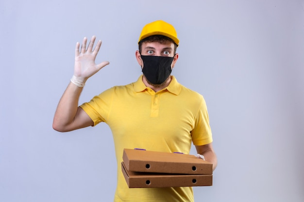 Livreur en polo jaune et casquette portant un masque de protection noir tenant des boîtes de pizza montrant le numéro cinq avec la main à la surprise debout sur blanc