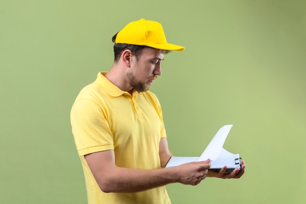 Livreur en polo jaune et casquette à la recherche de blancs dans sa main avec un visage sérieux debout sur vert isolé