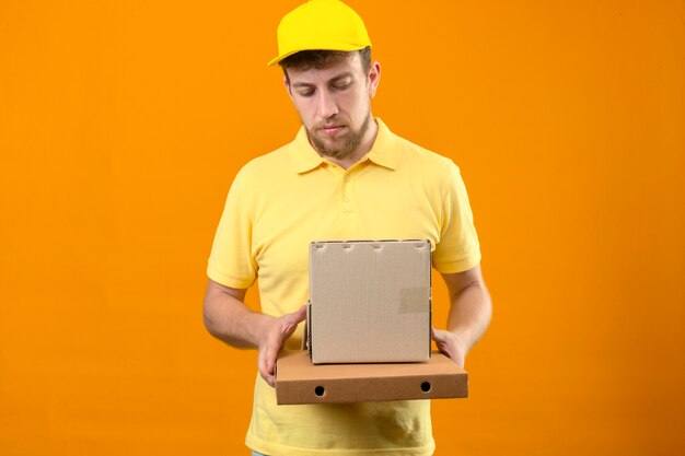 Livreur en polo jaune et casquette tenant des boîtes en carton à la triste avec visage malheureux debout sur orange isolé