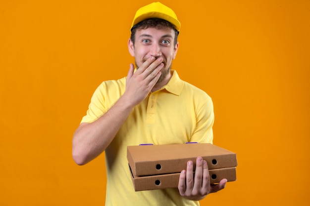 Livreur en polo jaune et casquette tenant des boîtes de pizza à la bouche coning surpris avec la main debout sur orange isolé