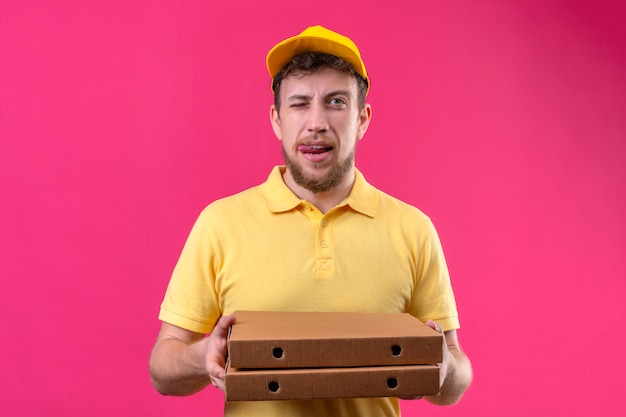 Livreur en polo jaune et casquette tenant des boîtes de pizza à un clin de œil joyeux sticking out tongue debout sur rose isolé