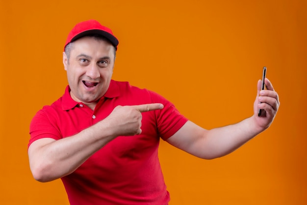 Livreur portant un uniforme rouge et une casquette à la joyeuse holding smartphone et pointant avec l'index vers lui souriant debout