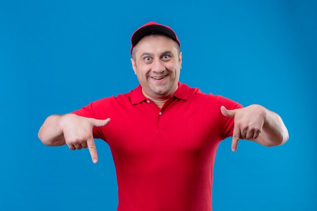 Livreur portant un uniforme rouge et une casquette à la surprise et heureux pointant vers le bas avec les doigts sur le mur bleu