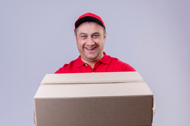 Livreur portant l'uniforme rouge et casquette tenant une grande boîte en carton souriant amical avec visage heureux sur mur blanc isolé