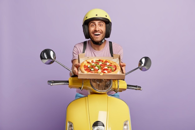 Photo gratuite livreur à la recherche amicale conduisant un scooter jaune tout en tenant la boîte à pizza