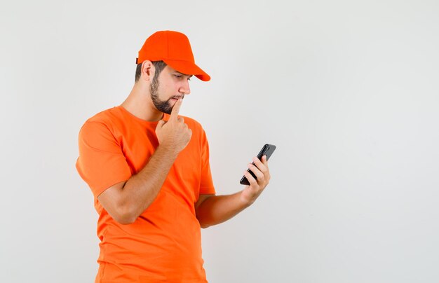 Livreur regardant un téléphone portable en t-shirt orange, casquette et semblant pensif. vue de face.