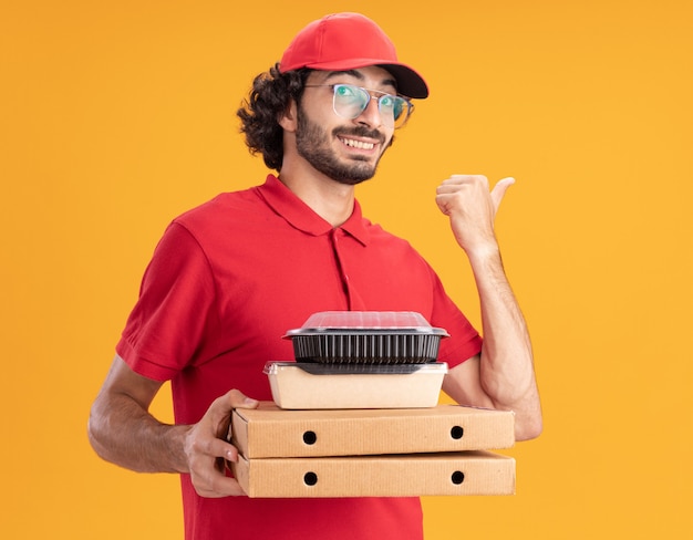 Photo gratuite livreur souriant en uniforme rouge et casquette portant des lunettes tenant des emballages de pizza avec un emballage de nourriture en papier et un récipient de nourriture sur eux regardant l'avant pointant sur le côté