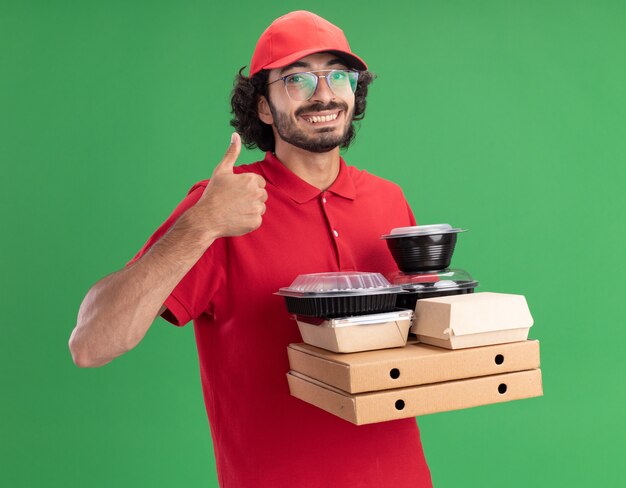 livreur souriant en uniforme rouge et casquette portant des lunettes tenant des emballages de pizza avec des emballages de nourriture en papier et des contenants de nourriture sur eux regardant l'avant montrant le pouce vers le haut