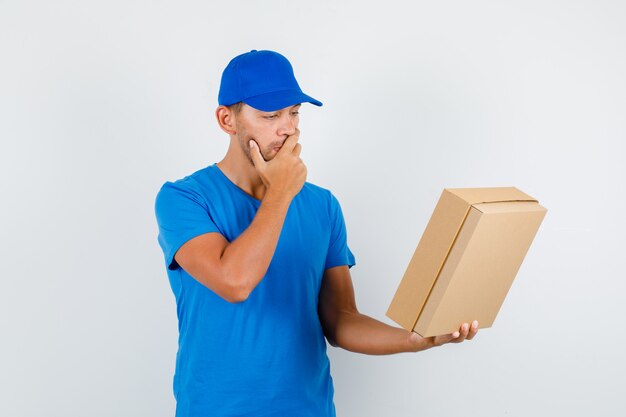 Livreur en t-shirt bleu, casquette tenant une boîte en carton avec la main sur la bouche et à la pensif