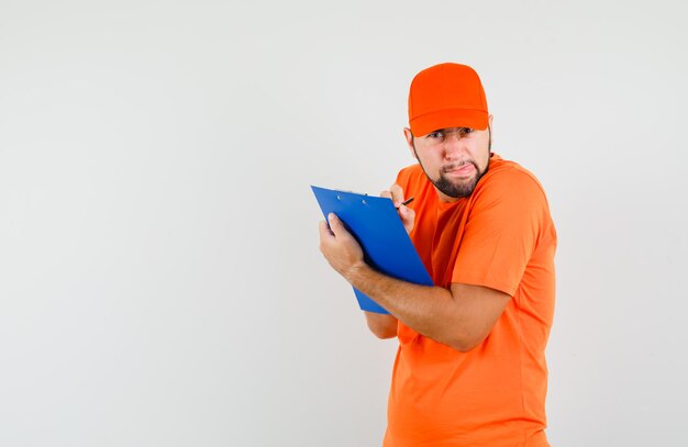 Livreur en t-shirt orange, casquette prenant des notes sur le presse-papiers et ayant l'air bizarre, vue de face.