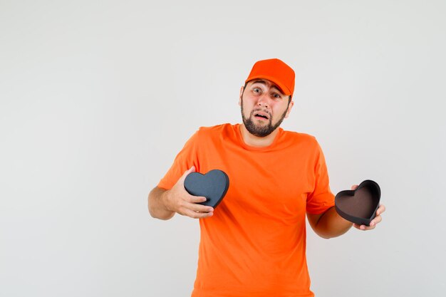 Livreur tenant une boîte-cadeau ouverte en t-shirt orange, casquette et regardant abattu, vue de face.