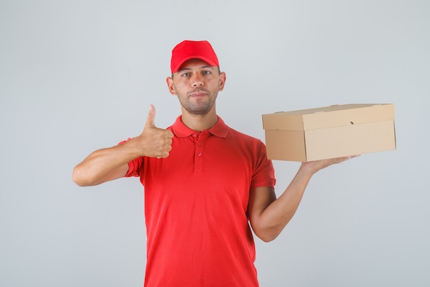 Livreur tenant une boîte en carton et montrant le pouce vers le haut en uniforme rouge et à la recherche positive.