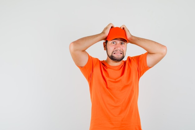 Livreur tenant les mains sur la tête en t-shirt orange, casquette et l'air impuissant. vue de face.