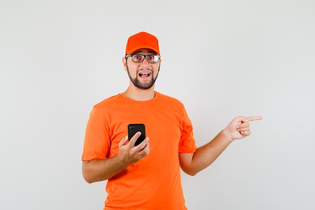 Livreur tenant un téléphone portable et pointant sur le côté en t-shirt orange, casquette, vue de face.