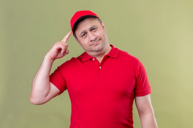 Livreur en uniforme rouge et cap pointant le temple avec le doigt se concentrant dur sur une idée debout sur le vert