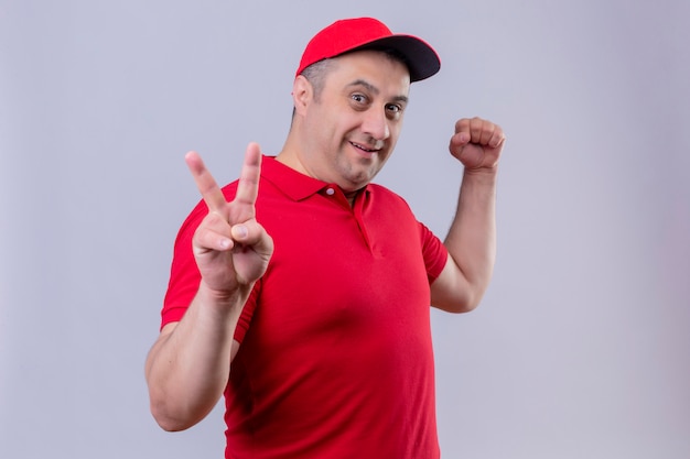 Livreur en uniforme rouge et cap à la recherche positive et heureuse souriant joyeusement montrant signe de la victoire avec deux doigts debout sur blanc isolé