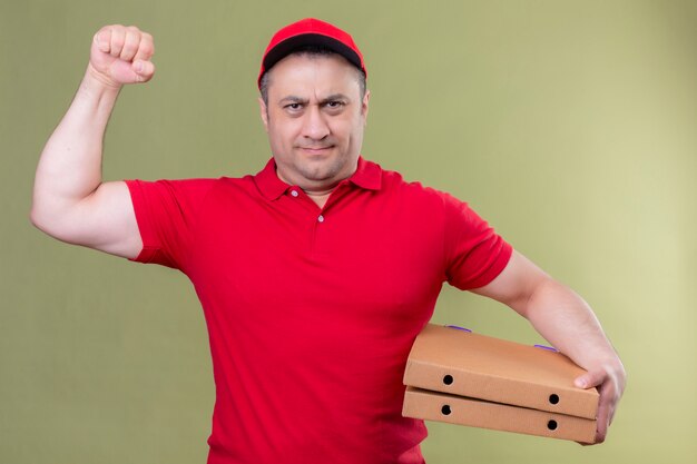 Livreur en uniforme rouge et cap tenant des boîtes à pizza montrant les biceps sur sa main, souriant confiant, concept gagnant debout sur le vert