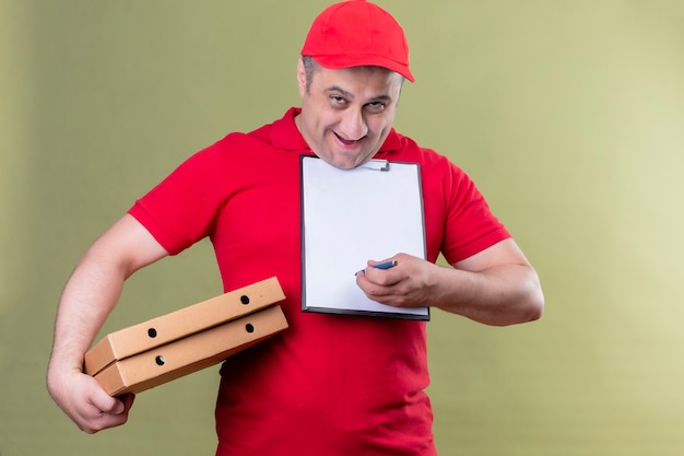 Photo gratuite livreur en uniforme rouge et cap tenant des boîtes à pizza et presse-papiers avec des blancs demandant la signature souriant sympathique debout sur l'espace vert