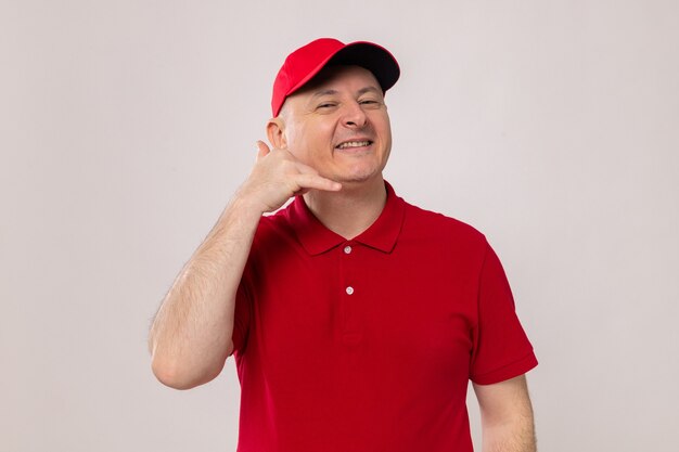 Livreur en uniforme rouge et casquette regardant la caméra souriant joyeusement me faisant appeler le geste debout sur fond blanc