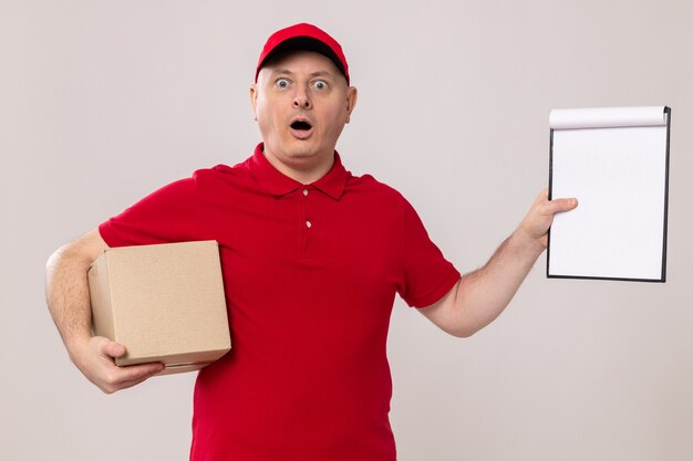 Livreur en uniforme rouge et casquette tenant une boîte en carton et un presse-papiers avec des pages blanches l'air étonné et surpris