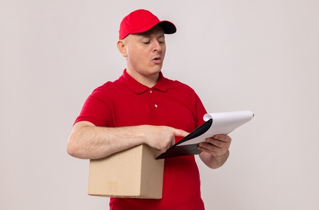 Livreur en uniforme rouge et casquette tenant une boîte en carton et un presse-papiers le regardant avec un visage sérieux debout sur fond blanc