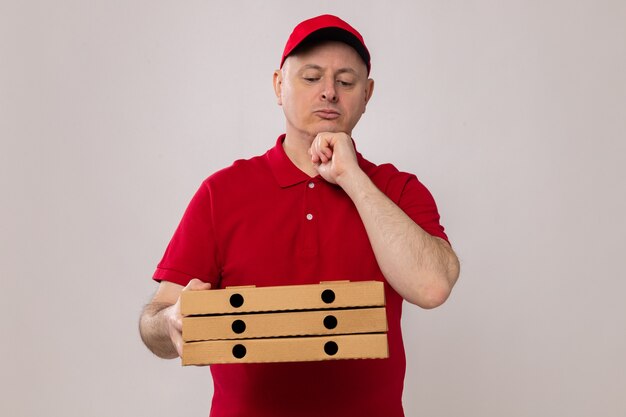 Livreur en uniforme rouge et casquette tenant des boîtes à pizza en les regardant avec une expression pensive sur le visage pensant debout sur fond blanc