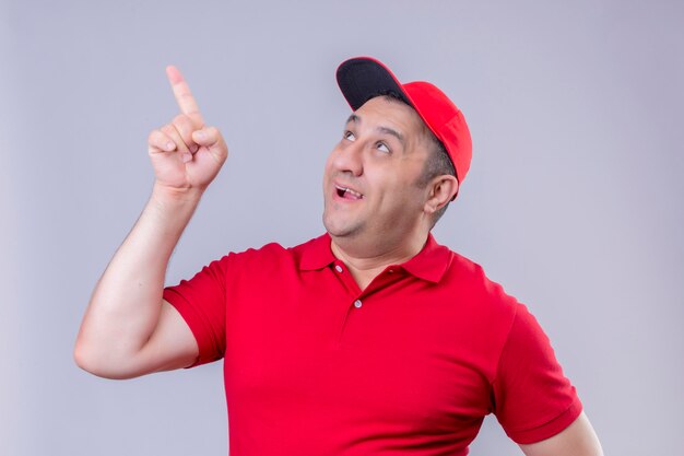 Livreur en uniforme rouge et chapeau souriant à la recherche et pointant du doigt quelque chose debout sur blanc