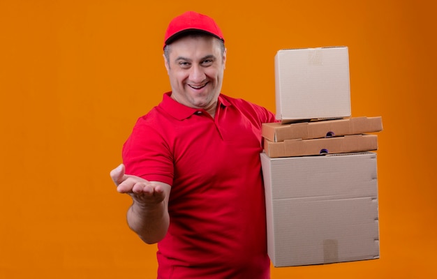 Livreur vêtu d'un uniforme rouge et casquette tenant des boîtes en carton à la recherche positive et heureuse de pointage avec le bras oh part à la caméra sur mur orange isolé