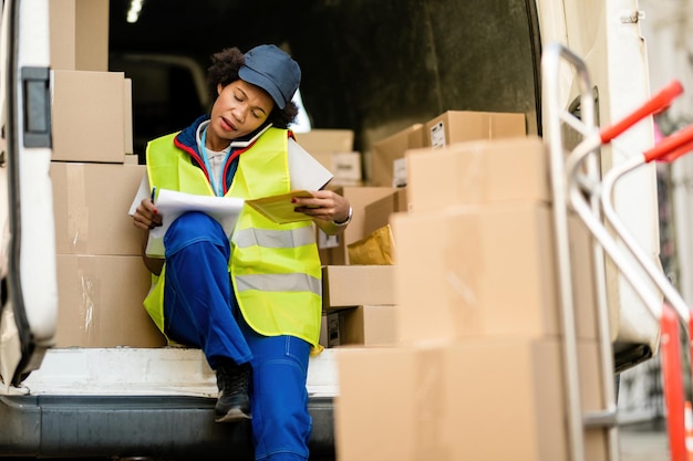 Photo gratuite livreuse afro-américaine parlant au téléphone tout en parcourant la liste de livraison dans une camionnette