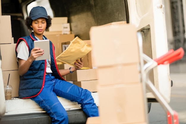 Livreuse noire parcourant la liste de contrôle sur le pavé tactile tout en vérifiant le colis de livraison dans un camion