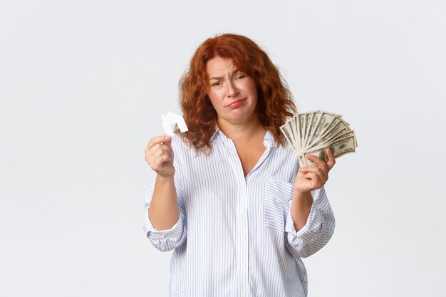 Location, achat de propriété et concept immobilier. Femme d'âge moyen rousse non amusée et triste tenant de l'argent et une petite carte de la maison, n'a pas assez d'argent, a besoin d'un prêt pour l'achat, mur blanc.