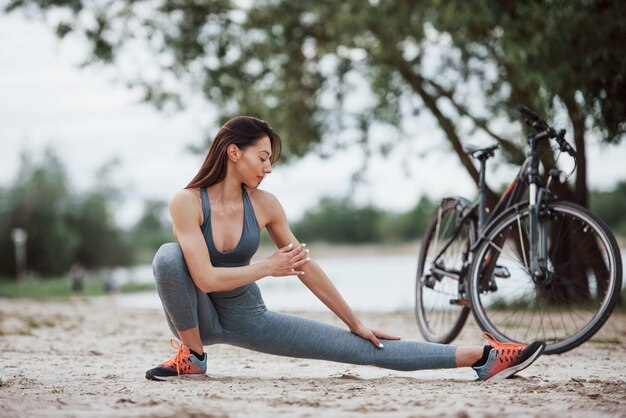 Look concentré. Cycliste féminine avec une bonne forme de corps faisant des exercices de yoga et des étirements près de son vélo sur la plage pendant la journée