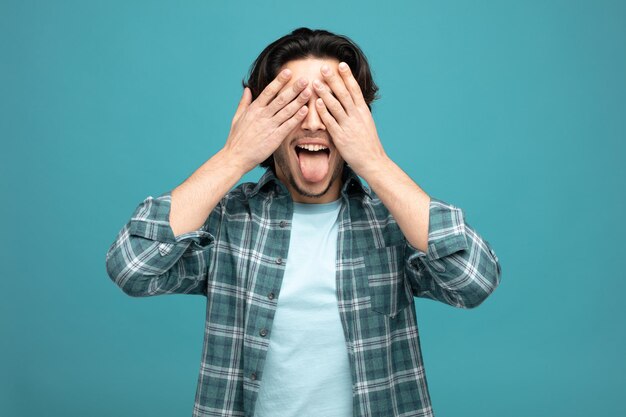 ludique jeune bel homme fermant les yeux avec les mains montrant la langue isolé sur fond bleu