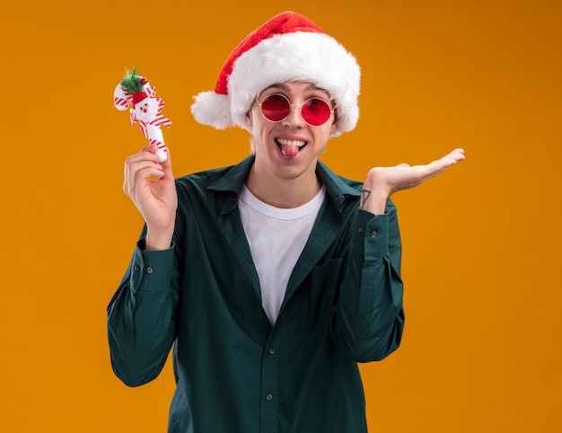 Ludique jeune homme blond portant bonnet de Noel et lunettes tenant l'ornement de canne douce regardant la caméra montrant la langue et la main vide isolée sur fond orange