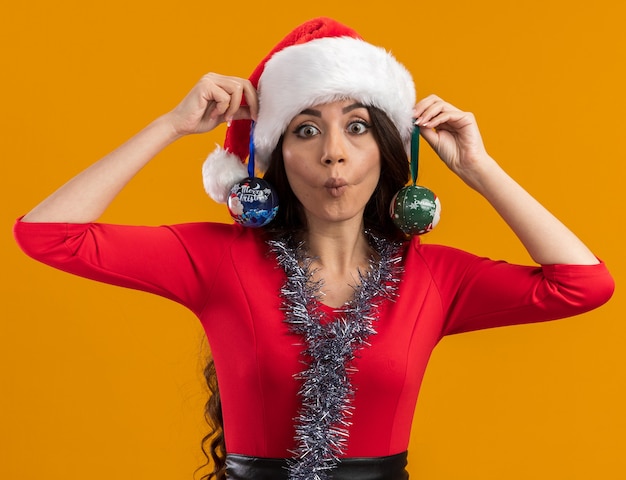 Ludique jeune jolie fille portant bonnet de Noel et guirlande de guirlandes autour du cou suspendus boules de Noël sur les oreilles regardant la caméra faisant face de poisson isolé sur fond orange