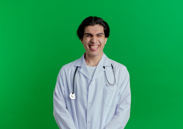Ludique jeune médecin de sexe masculin portant une robe médicale et un stéthoscope montrant la langue isolée sur un mur vert avec espace copie
